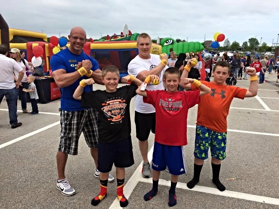 Strongmen and kids flexing in front of bounce houses at outdoor event